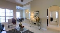This single-story residence is designed with an open-concept layout that effortlessly connects the living, dining, and kitchen areas into one cohesive and welcoming space. The living area features light-toned carpet, a glass-top coffee table, and a ceiling fan overhead, creating a comfortable and functional gathering space. A sleek chrome console table adorned with decorative vases and artwork adds a refined touch to the transitional zone between rooms.

The dining area accommodates a full table and chair set beneath a ceiling-mounted track lighting fixture, providing ample illumination for mealtimes. Natural light filters in through a large window, brightening the space and enhancing the airy feel created by the notably high ceilings that run throughout.

The kitchen is partially open to the main living area, separated by a raised breakfast bar with a granite or stone-look countertop. Pendant lights hang above the bar, adding warmth and a modern aesthetic. A white panel door likely leads to a closet or utility space, contributing to the home's clean and organized design.

The soft neutral color palette of warm beige and white on the walls and flooring unifies all areas, lending the home a calm and inviting ambiance. The thoughtful layout and elevated ceiling height give the entire space a generous sense of openness and scale.