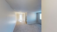 This single-floor space presents a clean, open layout characterized by neutral gray and beige tones throughout. The main living area is finished with plush wall-to-wall carpeting in a light gray hue, contributing to a comfortable and cohesive aesthetic. Walls are painted in a soft, neutral gray tone, complemented by white baseboards that provide a clean contrast.

A textured popcorn ceiling runs throughout the space, adding a classic residential feel. Natural light enters through a window fitted with horizontal blinds, brightening the interior and enhancing the overall sense of openness. A baseboard heater is positioned along the exterior wall, providing an efficient heating solution.

A hallway connects to additional areas of the unit, leading toward what appears to be a well-lit room featuring a modest chandelier, suggesting a dining or entry area. The corridor also reveals a doorway and what may be additional closet or utility access.

The overall ambiance is calm and understated, with the neutral palette and simple finishes creating a versatile canvas well-suited for a variety of interior styles.