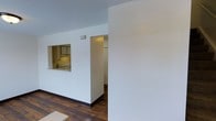 This two-floor space offers a functional and straightforward layout designed for comfortable everyday living. The main level features rich, dark hardwood floors that contrast cleanly against bright white walls, creating a classic and timeless aesthetic throughout. A notable architectural detail is the pass-through window cut into the interior wall, which connects the living area to the kitchen — promoting an open feel while maintaining defined zones. The kitchen is equipped with standard cabinetry and appliances, offering practical functionality in a compact footprint. Adjacent to the kitchen, a doorway leads to an additional room, likely a utility or storage space, visible through a set of louvered doors. The clean, minimal design of the space allows for versatile furnishing arrangements. White walls and neutral tones throughout contribute to a bright, airy ambiance, while the dark hardwood flooring adds warmth and character. With two floors, the space provides ample room for separation between living and private areas. Overall, the layout balances openness with defined spaces, making it a practical and adaptable environment.