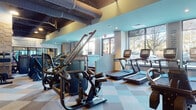 This contemporary fitness facility is thoughtfully designed to accommodate a full spectrum of workout needs across a single expansive floor. The space features a striking industrial-chic aesthetic, with exposed ductwork and dark ceiling beams contrasting against teal accent walls, stone-clad columns, and a multi-toned checkered rubber tile flooring that defines each training zone with visual clarity.

The cardio section lines the perimeter wall of large windows, offering an impressive row of treadmills and elliptical machines bathed in natural light with outdoor views of surrounding greenery. Adjacent to this is a dedicated strength and power zone, housing an extensive dumbbell rack, adjustable benches, and a variety of cable and selectorized machines. A boxing bag and motivational wall typography — including phrases like "The Best Investment Is Your Own Health" and "Opportunities Don't Happen, You Create Them" — add an energizing atmosphere throughout.

A separate functional training area features BOSU balls, open floor space with wood-look flooring, mirrored walls, and a stair climber machine. A welcoming entry corridor with framed geometric artwork and glass-panel doors leads seamlessly into the main floor. The facility's layout is open, airy, and well-lit, making it a dynamic and inspiring environment for all fitness levels.