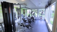 This fitness room is designed to support a well-rounded workout experience within a compact, efficiently arranged single-floor layout. The space features durable rubber flooring, which provides both safety and sound absorption during exercise. Large windows along one wall flood the room with natural light and offer pleasant views of surrounding greenery, creating an open and energizing atmosphere.

The room is equipped with a range of fitness equipment, including a multi-function cable weight machine, an elliptical trainer, and a treadmill, catering to both strength and cardio training needs. A medicine ball rack holding multiple weighted balls of varying sizes is positioned near the windows, adding versatility for functional fitness routines. Full-length mirrors are mounted along one wall, allowing users to monitor their form during workouts and visually expanding the sense of space.

The walls are painted in a clean white with green accent stripes, giving the room a fresh, energetic aesthetic. Recessed and surface-mounted lighting complements the natural light from the windows. The overall design is practical and functional, offering a motivating environment for daily fitness routines.