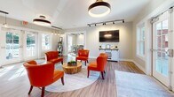 Located at 4122 Laurel Village Dr in Henrico, Virginia, this single-story community clubhouse offers a well-appointed gathering space designed for both relaxation and productivity. The interior is finished with warm hardwood-style flooring that flows consistently throughout, complementing the neutral gray-toned walls and crisp white trim.

The central lounge area serves as the heart of the space, furnished with bold orange leather accent chairs arranged around a natural wood stump coffee table. A wall-mounted flat-screen television and a white barn-style console anchor one side of the room, while large French doors with multi-pane windows flood the area with natural light and provide views of the surrounding outdoor landscape. Overhead, statement ring pendant light fixtures add a modern touch alongside adjustable track lighting.

Adjacent to the lounge, a dedicated workspace features a conference-style table, task seating, pendant lighting, and glass-panel partition doors, creating a semi-private environment suited for meetings or focused work.

The kitchen area is outfitted with white shaker cabinetry, granite countertops, under-cabinet LED lighting, stainless steel appliances, and a single-basin undermount sink, offering practical amenities for community gatherings. Overall, the space presents a clean, contemporary aesthetic with inviting design details.