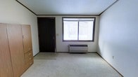 This single-floor space offers a straightforward and functional layout designed for comfortable living. The main room is finished with light-colored wall-to-wall carpeting that adds warmth underfoot, while white walls and a textured ceiling contribute to a clean, neutral aesthetic throughout. A large window with a wood-trimmed frame allows an ample amount of natural light to fill the room, offering views of the surrounding outdoor environment. Beneath the window sits a wall-mounted HVAC unit, providing convenient climate control directly within the living space. A dark-stained solid door with a modern lever handle serves as the primary entry point, providing a subtle contrast against the lighter tones of the room. Crown molding and baseboards with a dark wood finish frame the walls, adding a touch of architectural detail and definition to the space. The room's open, uncluttered layout offers flexibility for a variety of furniture arrangements. Overall, the space presents a modest yet practical environment with neutral finishes that provide a versatile backdrop for personalization.