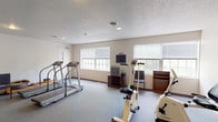 The fitness center at 2801 Montaluma Dr in Springfield, Illinois, is a dedicated exercise facility housed on a single floor. The space features a practical and functional layout designed to accommodate a variety of cardio-focused workouts. The room is finished with durable wall-to-wall carpeting in a neutral gray tone, complemented by clean white walls and a textured ceiling with recessed lighting that provides consistent, even illumination throughout.

Multiple treadmills are positioned along one side of the room, offering ample space for comfortable use. A stationary upright bike and an arm cycle station are also available, providing a range of low-impact cardiovascular options. A rowing machine rounds out the cardio equipment selection. A small table with chairs offers a place to rest between sessions, and a wall clock keeps users on schedule.

Natural light flows in through several large windows fitted with horizontal blinds, giving the space an open and airy feel. A television and media console are stationed along one wall, allowing users to watch programming during their workouts. The facility is accessible via a double-door exit and a standard entry door. Overall, the fitness center offers a straightforward, well-equipped environment suited for residents seeking convenient access to regular exercise.