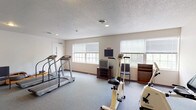 The fitness center at 2801 Montaluma Dr in Springfield, Illinois, is a dedicated exercise facility housed on a single floor. The space features a practical and functional layout designed to accommodate a variety of cardio-focused workouts. The room is finished with durable wall-to-wall carpeting in a neutral gray tone, complemented by clean white walls and a textured ceiling with recessed lighting that provides consistent, even illumination throughout.

Multiple treadmills are positioned along one side of the room, offering ample space for comfortable use. A stationary upright bike and an arm cycle station are also available, providing a range of low-impact cardiovascular options. A rowing machine rounds out the cardio equipment selection. A small table with chairs offers a place to rest between sessions, and a wall clock keeps users on schedule.

Natural light flows in through several large windows fitted with horizontal blinds, giving the space an open and airy feel. A television and media console are stationed along one wall, allowing users to watch programming during their workouts. The facility is accessible via a double-door exit and a standard entry door. Overall, the fitness center offers a straightforward, well-equipped environment suited for residents seeking convenient access to regular exercise.