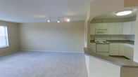 This single-floor residential space features an open-concept design that seamlessly connects the living area and kitchen. The living room is finished with neutral-toned wall-to-wall carpeting and painted in a soft, light gray that contributes to a clean, understated aesthetic. A track lighting fixture mounted on the ceiling provides adjustable illumination throughout the space, complementing the textured ceiling above.

The kitchen is accessible via an arched passageway and is partially open to the living area through a raised breakfast bar counter finished in a warm neutral tone, offering a functional divide between the two spaces. The kitchen is equipped with white cabinetry providing ample storage, a built-in microwave, and an electric range with overhead ventilation, all maintaining a cohesive white appliance scheme. The kitchen counter space is practical and compact, suitable for everyday meal preparation.

Overall, the space presents a functional and straightforward layout with a neutral color palette throughout, making it adaptable to a variety of furnishing styles. The open sightlines between the living and kitchen areas enhance the sense of space and connectivity within the unit.