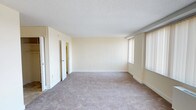 This single-floor space presents a generously sized main living area finished with neutral, cream-colored walls that contribute to a bright and airy atmosphere. The room is covered in wall-to-wall carpet in a neutral beige tone, providing a soft and comfortable underfoot feel throughout. A textured popcorn ceiling adds a classic finish overhead, while clean white baseboards frame the perimeter of the room neatly.

Natural light flows into the space through multiple windows fitted with vertical blinds, allowing for adjustable light control. The windows are positioned to brighten the room effectively, enhancing its open and welcoming feel.

Toward the rear of the main area, two doorways lead to additional rooms or spaces beyond, suggesting further utility areas such as a hallway, closets, or additional rooms. The layout appears straightforward and functional, with an open floor plan that allows for flexible furniture arrangement.

Overall, the space carries a clean, understated aesthetic with its consistent neutral palette, ample square footage, and practical finishes, making it a versatile and livable environment.