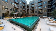 This contemporary amenity space is defined by thoughtful design and high-quality finishes that create an inviting, functional environment. At the heart of the property lies a stunning courtyard pool with vivid turquoise water, framed by natural stone river rock borders and surrounded by a spacious pool deck lined with lounge chairs and shaded umbrella seating. The pool is set within a multi-story building courtyard, with private balconies overlooking the water on all sides, creating a resort-like atmosphere with a strong sense of enclosure and community.

Inside, the space includes a well-appointed conference room that combines modern sophistication with creative flair. Bold color-blocked wall panels in yellow, gray, and white serve as a striking backdrop, complemented by a wall-mounted display screen and a large whiteboard. A sleek wood-topped conference table is centered in the room beneath a sculptural gold geometric pendant light that serves as a dramatic focal point. Glass partition walls with sheer drapery panels divide the interior spaces, allowing natural light to filter through while maintaining a sense of openness. Recessed lighting and stone-look tile wall finishes add to the polished, contemporary aesthetic throughout, making this amenity space both visually impressive and highly functional.