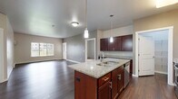 This two-bedroom, two-bathroom unit in West Fargo, North Dakota offers a thoughtfully designed single-floor layout with a comfortable and functional living environment. The open-concept kitchen and living area serves as the heart of the home, anchored by a central island topped with granite countertops, rich dark-stained cabinetry, pendant lighting, and stainless steel appliances. Dark hardwood flooring flows throughout the main living areas, complementing the neutral taupe and gray wall tones that lend a clean, modern feel. The living room receives natural light through large windows fitted with vertical blinds, and features a private entry door. Both bedrooms are carpeted and generously sized, offering a quiet retreat with ample natural light. The primary bathroom includes granite-topped vanity cabinetry matching the kitchen aesthetic, dark hardwood floors, and direct access to a dedicated walk-in closet with built-in shelving. The second bathroom mirrors the same finishes and includes a full tub-shower combination. A dedicated in-unit laundry area accommodates a full-size washer and dryer side by side, with an overhead shelf for added storage. Two closets throughout the unit provide practical organization options for residents.