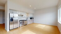 This single-floor residence offers a clean, contemporary design characterized by an all-white color palette, light hardwood flooring, and recessed lighting that creates a bright and airy atmosphere throughout. The open-concept kitchen is a standout feature, outfitted with stainless steel appliances including a refrigerator, range, dishwasher, and microwave. White upper cabinetry contrasts with light wood-toned lower cabinets, while a glossy white tile backsplash adds a polished finishing touch. Ample counter space and generous cabinetry provide excellent storage and functionality. A hallway connects the living areas to the private spaces, maintaining a logical and efficient floor plan. The bedroom includes a sliding-door closet with built-in shelving and benefits from natural light through a well-positioned window. The full bathroom is finished with hexagonal floor tiles, a wood-toned vanity with an integrated sink, a large mirror, and a soaking tub with a shower combination. A dedicated in-unit laundry area with a washer and dryer is discreetly tucked behind a sliding door, adding everyday convenience. Overall, the space delivers a modern, functional living environment with thoughtful finishes and a cohesive, minimalist aesthetic.