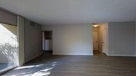This single-floor unit offers a straightforward, functional layout designed for comfortable everyday living. The main living area is spacious and open, finished with smooth hardwood-style flooring that extends throughout the space, providing a clean and modern aesthetic. Neutral grey walls and white baseboards create a calm, versatile backdrop suitable for a variety of interior styles. The textured ceiling adds a subtle architectural detail overhead. Natural light filters into the living area, casting soft patterns across the floor and contributing to a bright, welcoming atmosphere. Visible from the main living space is a connected kitchen area, which features white appliances and a built-in shelving nook, offering practical storage solutions. An entryway is positioned to one side, framed by a white door and a lit overhead fixture, providing a clearly defined transition between the interior and exterior. A visible HVAC vent integrated into the wall ensures year-round climate control. The overall layout is efficient and cohesive, with each area flowing naturally into the next, making the space easy to navigate and adaptable to various living arrangements.