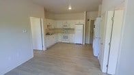This single-floor space offers a clean, functional layout centered around an open-concept kitchen and living area. The kitchen is well-appointed with white cabinetry providing ample storage, a full suite of white appliances including a refrigerator, electric range, and over-the-range microwave, as well as a sink integrated into the countertop along the wall. The cabinetry lines two walls in an L-shaped configuration, maximizing workspace and storage efficiency. Light wood-look flooring extends throughout the space, lending a bright and airy atmosphere that complements the neutral, light-toned walls. Recessed lighting in the ceiling provides consistent illumination across the open area. The generous open floor plan between the kitchen and the adjacent living space allows for flexible furniture arrangements and comfortable movement throughout. Multiple doorways provide access to additional areas of the unit, contributing to a practical and connected flow. The overall aesthetic is crisp and minimal, with a consistent white-and-neutral palette that creates a welcoming, move-in-ready environment suitable for a variety of uses.