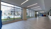 This single-floor fitness studio is designed with a clean, contemporary aesthetic that prioritizes openness and natural light. Expansive floor-to-ceiling windows line the exterior walls, offering unobstructed views of an outdoor pool area complete with lounge seating and manicured green space, seamlessly connecting the interior to the surrounding courtyard environment.

The space features smooth light-toned hardwood-style flooring throughout, providing an ideal surface for yoga, stretching, or group fitness activities. Recessed linear LED lighting runs across the ceiling in parallel lines, delivering consistent, even illumination that complements the abundant natural light from the large window panels.

A full-length mirror is positioned along one wall, enhancing the sense of space while supporting functional fitness use. A dedicated equipment rack, stocked with resistance and training tools, is visible toward the rear of the room, indicating a dual-purpose layout suitable for both open floor exercise and structured workouts.

The neutral color palette of warm beige walls and light flooring contributes to a calm, focused atmosphere, making this studio a well-appointed and versatile fitness amenity.