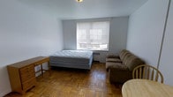 This single-floor studio unit offers a straightforward and functional open-plan layout that combines sleeping, living, and relaxing areas within one unified space. The room is finished with warm-toned classic parquet hardwood flooring arranged in a herringbone pattern, adding a timeless character to the interior. A centrally positioned window fitted with horizontal blinds allows natural light to filter into the space while providing privacy control. Beneath the window sits a wall-mounted HVAC unit, ensuring year-round climate comfort. The room accommodates a full-sized bed, a sofa, and a small side table, demonstrating how the space can be efficiently arranged for both rest and leisure. The ceiling features a flush-mounted light fixture that provides overhead illumination. The walls are painted in a clean, neutral light gray tone, lending a calm and airy ambiance to the space. The overall layout is simple and adaptable, making it well-suited for a single occupant seeking a no-frills, self-contained living environment. The minimalist design keeps the focus on functionality and ease of use throughout.