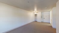 This single-floor unit offers a practical and functional layout across multiple rooms, all finished with neutral tones and wall-to-wall carpeting throughout the living and sleeping areas. The main living space is generously proportioned, featuring a ceiling fan with light fixture, a window overlooking surrounding greenery, and a wall-mounted HVAC unit providing climate control. The textured ceiling adds subtle detail to the room.

The bedroom areas are well-sized, each carpeted and finished with clean white walls. One bedroom includes a reach-in closet and ceiling fan, while another features a large window with a baseboard heater beneath it, allowing natural light and views of mature trees outside. An additional room provides flexible use as a bedroom or office, with neutral carpeting and ample wall space.

The galley-style kitchen is efficiently designed with light wood cabinetry lining both sides, offering substantial storage. White appliances include a coil-burner range, refrigerator, and dishwasher, complemented by a single-basin sink and light-toned countertops. The floor is finished with vinyl tile.

The bathroom features a full tub-shower combination equipped with multiple stainless steel grab bars for accessibility, along with a handheld showerhead. Built-in open shelving provides convenient storage, and a vanity with wood cabinetry completes the space. The floor is finished with patterned vinyl tile.