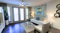 This single-floor space combines modern design with comfortable living. The living area serves as the centerpiece of the home, showcasing rich, dark hardwood flooring that extends throughout the space and lends a warm, polished aesthetic. A ceiling fan with integrated lighting anchors the room from above, while a set of French doors flanked by wide windows invites an abundance of natural light, creating an airy and open atmosphere. Elegant floor-length drapes complement the blinds, offering both style and privacy. The living area is thoughtfully appointed with a crisp white sofa adorned with teal and patterned accent pillows, paired with a matching white armchair and a sleek glass and chrome coffee table atop a plush area rug. A large, framed teal and silver wall art piece serves as a striking focal point, adding a vibrant yet sophisticated touch to the neutral palette. A classic table lamp provides warm ambient lighting, enhancing the inviting atmosphere of the room. The overall ambiance of the space is contemporary and well-curated, blending clean lines with carefully chosen accents to create a comfortable and visually appealing environment.