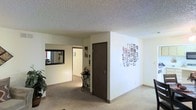 This single-story space presents an open and functional layout characterized by clean white walls and neutral-toned carpeting that flows throughout the interior. The textured popcorn ceiling adds a classic architectural detail overhead, lending a familiar, understated aesthetic to the space.

A central interior door, finished in a dark brown tone, provides access to an adjoining room or private area, adding a contrast against the light-colored walls. A passageway visible in the background leads further into the space, suggesting a connected layout with multiple areas accessible from the main living zone.

A framed wall mirror is mounted near the passageway, enhancing the sense of depth and light within the space. Beneath it, a small decorative accent table with an ornamental figurine adds a subtle design element. On the opposite wall, a collage-style piece of decorative wall art serves as a focal point, introducing visual interest to the otherwise minimalist aesthetic.

Natural light filters in through a window visible in the background, contributing to the bright, open feel of the space. Overall, the interior conveys a straightforward, livable environment with a neutral palette and simple, functional design elements.