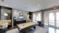 This single-floor recreational space is thoughtfully designed to blend entertainment and comfort in a contemporary setting. At its center sits a full-size billiards table positioned atop a neutral area rug, serving as the focal point of the room. Recessed lighting in the smooth white ceiling provides consistent illumination throughout the space.

An accent wall clad in stacked stone tile houses a large flat-screen television alongside additional mounted displays, creating a dedicated media zone. Dark cabinetry beneath the screens offers storage and counter space, accompanied by what appears to be a beverage station, adding functional convenience to the entertainment area.

Adjacent to the media wall, a cozy lounge nook features a pendant light fixture and comfortable seating, providing an intimate retreat within the larger space. The room transitions seamlessly toward a bright seating area positioned near expansive floor-to-ceiling windows dressed with sheer, striped curtains. These windows flood the space with natural light and offer views of an outdoor patio area. Dark hardwood-style flooring grounds the room, tying together the modern, sophisticated aesthetic throughout.