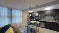 This single-floor residence showcases a thoughtfully designed open-concept layout that seamlessly connects the kitchen and dining area, creating an inviting and functional living environment. The kitchen is a standout feature, outfitted with dark espresso cabinetry accented by brushed nickel hardware, granite countertops, and a striking mosaic tile backsplash that adds texture and visual depth. Stainless steel appliances, including a refrigerator, electric range, microwave, and dishwasher, provide a polished, contemporary look. A deep stainless steel sink with a modern arc faucet anchors the countertop workspace. Flush-mount ceiling fixtures and recessed lighting illuminate the kitchen efficiently. The adjacent dining area is defined by an industrial-style bistro table with metal barstools, lit by stylish pendant lights that hang gracefully overhead. Light wood-tone flooring runs throughout, lending warmth and continuity to the space. Large windows dressed with sheer curtains and horizontal blinds flood the interior with natural light, enhancing the open and airy ambiance. The overall design strikes a balance between modern sophistication and everyday practicality, making this a comfortable and well-appointed living space.