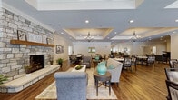 Located at 2832 Welcome Way in Evansville, Indiana, this single-story community facility is thoughtfully designed to offer a variety of shared spaces that blend comfort with refined aesthetics. Throughout the building, warm hardwood-style flooring, crown molding, and neutral tones create a cohesive and inviting atmosphere.

The central gathering area serves as the heart of the facility, anchored by a striking floor-to-ceiling stone fireplace surrounded by comfortable seating arrangements. Tray ceilings with elegant chandeliers add architectural depth, while the open layout flows seamlessly into a dining area furnished with multiple tables and chairs. A service counter connects to a kitchen pass-through, supporting dining operations efficiently.

A dedicated sitting area features upholstered armchairs alongside natural light from multiple windows, providing a quieter retreat. An adjacent room houses a grand piano set atop a patterned area rug, accompanied by framed artwork, making it well-suited for musical performances or entertainment.

A chapel and presentation room offers rows of padded seating, a projection screen, and a ceiling-mounted projector, ideal for gatherings or screenings. Additional spaces include a lounge with a wall-mounted television and a large, open multipurpose room with wainscoting and ample floor space for events or activities.