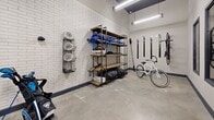 Located at 360 Acoma St in Denver, Colorado, this equipment room is a well-organized, single-level storage facility designed to accommodate a range of recreational and operational gear. The room features a practical, industrial aesthetic with polished concrete floors, white subway tile accent walls, and neutral painted surfaces throughout. Overhead fluorescent lighting ensures the space is well-lit, supporting easy navigation and inventory management.

The layout is divided into distinct functional zones. A primary storage area houses multiple freestanding industrial-style wood-and-metal shelving units, providing ample tiered storage for helmets, bags, coolers, and miscellaneous equipment. Wall-mounted wire basket organizers offer additional storage for smaller items. A dedicated bike storage section features vertical wall-mounted bike racks capable of holding several bicycles, keeping floor space clear and organized.

Access to the room is provided through sturdy charcoal-framed double doors with glass panels, which also connect to an adjacent corridor area where a mobile rolling cart serves as a staging surface. The room's design prioritizes functionality and durability, with heavy-duty finishes suited to frequent use. Overall, this equipment room presents a clean, efficient, and well-structured environment for centralized storage and equipment distribution.