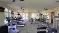 This fitness center offers a bright and open workout environment designed to accommodate a full range of exercise needs. The space is lined with large windows along multiple walls, flooding the interior with natural light and providing views of the surrounding outdoor area. Neutral-toned walls and patterned carpet flooring contribute to a clean, functional aesthetic throughout.

The cardio section is prominently featured, with a long row of treadmills and additional cardio machines, including stationary bikes, positioned to face the expansive windows. Strength training equipment, including cable machines and adjustable benches, is arranged throughout the remaining floor space, offering versatility for various workout routines.

Multiple wall-mounted flat-screen televisions are positioned throughout the room, ensuring visibility from different workout stations. Ceiling fans are evenly distributed across the ceiling, supplementing the climate control and maintaining air circulation throughout the space. Recessed lighting provides additional illumination during lower-light conditions. Full-length mirrors along one wall enhance the sense of openness while serving a practical function for form monitoring during exercise.