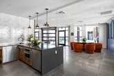 This thoughtfully designed single-floor amenity lounge offers a dynamic blend of modern and industrial aesthetics. The entry is marked by a striking blue patterned wallcovering accent wall and large glass doors that set an inviting tone. Inside, the open layout flows seamlessly across large-format tile flooring, connecting distinct seating and activity zones. A reclaimed wood feature wall serves as a dramatic focal point in the main lounge area, anchoring a flat-screen television and complementing the mix of plush gray swivel chairs, bold orange lounge seating, and organic-shaped wooden coffee tables atop vibrant blue area rugs. A dedicated dining and bar area is lined with floor-to-ceiling windows that flood the space with natural light and offer views of the surrounding cityscape. The kitchenette is outfitted with warm wood cabinetry, stainless steel appliances, a matte black faucet, and a glossy white subway tile backsplash, adding a refined yet functional touch. Pendant lighting with an industrial finish adds warmth overhead. The space also includes accessible restrooms finished with patterned tile flooring and blue-painted walls with gold-toned signage hardware. Throughout, the design balances comfort and character with curated artwork and lush greenery.