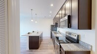 This thoughtfully designed 1-bedroom, 1-bathroom unit offers an efficient and modern living experience across a single floor. The kitchen is the standout feature, boasting rich dark-espresso cabinetry, granite countertops, and a full suite of stainless steel appliances including a refrigerator, electric range, microwave, and dishwasher. A central island provides additional prep space and casual seating, while pendant lighting overhead adds a warm, stylish accent. Light wood-style flooring runs throughout the kitchen and living areas, contributing to a bright and open ambiance. The bedroom is a comfortable retreat, carpeted for warmth and featuring a ceiling fan, a window with exterior views, and direct access to a dedicated walk-in closet complete with wire shelving and hanging rods. The bathroom continues the cohesive design language with dark cabinetry, granite countertops, and wood-style flooring. A hallway connects the living spaces to the private quarters, ensuring a logical and functional layout. Recessed lighting and clean white walls throughout the unit create a crisp, contemporary aesthetic that maximizes the sense of space.