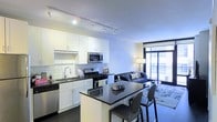 This stylish single-floor residence showcases a thoughtfully designed open-concept layout that seamlessly connects the kitchen and living areas. The kitchen is outfitted with stainless steel appliances, including a gas range, built-in microwave, and dishwasher, all complemented by crisp white cabinetry and a classic subway tile backsplash. Dark granite countertops provide striking contrast and extend to a convenient kitchen island with bar seating, making it an ideal space for both meal preparation and casual dining. Track lighting overhead ensures the kitchen is well-illuminated throughout. The living area flows naturally from the kitchen, offering a comfortable and inviting atmosphere anchored by a sophisticated tufted sofa and a patterned area rug over dark hardwood-style flooring. A floor-to-ceiling glass door and window wall at the far end of the living space flood the interior with natural light while providing direct access to a private balcony, offering views of the surrounding exterior. The overall ambiance balances modern elegance with everyday functionality, characterized by a clean, neutral color palette, high-quality materials, and a cohesive design aesthetic throughout.
