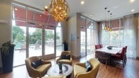 This single-floor space serves as an elegant communal lounge, designed to blend artistic flair with functional comfort. The room is anchored by a striking sculptural chandelier crafted from gold-toned lettering and symbols, creating an immediate focal point upon entry. Hardwood flooring runs throughout, lending warmth and continuity to the space.

Floor-to-ceiling glass windows and doors line the walls, flooding the interior with abundant natural light while offering unobstructed views of a landscaped outdoor pool area. The seamless indoor-outdoor connection is further emphasized by glass-paneled doors that open directly to the pool deck, where lounge chairs are visible.

The seating arrangement features plush, gold-toned armchairs with dark accent cushions arranged around a decorative round coffee table, creating an inviting conversational setting. A dining area with dark wooden chairs and a white table is positioned near the windows. Pendant lighting supplements the overhead illumination in the dining zone. The overall ambiance is sophisticated and welcoming, balancing modern design elements with comfortable furnishings suited for social gatherings or relaxed lounging.