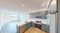 Located at 17111 Von Karman Ave in Irvine, California, this modern single-floor residence showcases a clean, contemporary design highlighted by light hardwood-style flooring that flows consistently throughout the space. The open-concept kitchen is a standout feature, equipped with sleek blue-gray flat-front cabinetry, a marble-look quartz countertop, and a generous island with a built-in sink. A full suite of stainless steel appliances — including a gas range, built-in microwave, dishwasher, and a French door refrigerator — provides a highly functional cooking environment. Elegant pendant lighting hangs above the island, complementing the recessed lighting overhead. The living area benefits from an abundance of natural light streaming through large sliding windows, with a glass door offering access to a private balcony and views of the surrounding Irvine cityscape. The bedrooms are bright and airy, each featuring ceiling fans, large windows with outdoor views, and ample wall space. The bathroom is finished with a blue-gray vanity, an illuminated mirror, built-in open shelving, and a tiled soaking tub and shower combination, all set against a clean, neutral palette that gives the space a fresh, modern feel throughout.