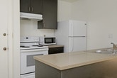 This single-floor space offers a clean, minimalist aesthetic defined by light hardwood-style flooring, crisp white walls, and a textured ceiling. The main living area is well-proportioned, featuring a ceiling fan with a built-in light fixture that adds both function and airflow. A set of built-in open shelves lines one wall, providing convenient storage and display space without compromising the room's open feel. A large window fitted with vertical blinds allows abundant natural light to filter in, casting dynamic patterns across the floor and contributing to the space's airy atmosphere. An open doorway connects the main living area to an adjacent room, which also benefits from natural light, suggesting a similarly bright and open layout throughout. The overall ambiance is neutral and versatile, well-suited for a variety of furniture arrangements and interior styles. The combination of natural light, clean finishes, and thoughtful built-in features makes this a functional and inviting single-level space.