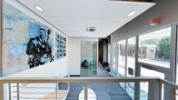 Located at 1202 Shelby St in Indianapolis' vibrant Fletcher Place neighborhood, the Forte Apartments leasing office is a thoughtfully designed single-floor space that blends contemporary architecture with creative character. The interior immediately draws attention with bold, music-themed wall art — including a striking large-scale portrait of a guitarist alongside an array of vinyl records — nodding to the building's artistic identity.

The space features a clean, modern aesthetic with a dropped grid ceiling, recessed lighting, and sleek white walls that keep the atmosphere bright and professional. A glass-partitioned private office provides a dedicated workspace while maintaining an open, transparent feel throughout. Floor-to-ceiling windows along the exterior wall flood the space with natural light and offer clear views of the surrounding streetscape, creating a welcoming and connected atmosphere.

A mezzanine-style overlook with a contemporary metal and cable railing provides views down to the lower level, adding a sense of vertical dimension and architectural interest. The flooring transitions between warm wood tones and plush blue carpet, complementing the cool, modern color palette. Comfortable seating areas are thoughtfully placed to accommodate visitors. Overall, the space exudes a polished yet inviting ambiance that reflects the energy and character of the Forte Apartments community.