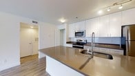 This single-floor space showcases a thoughtfully designed, contemporary kitchen that serves as its centerpiece. The kitchen is anchored by a generous island with a sleek, dark quartz countertop and an integrated sink fitted with a professional-style pull-down faucet, providing ample workspace and functionality. White flat-panel cabinetry lines the walls, extending to the ceiling to maximize storage, while a classic white subway tile backsplash adds a clean, timeless detail along the cooking wall. Stainless steel appliances, including an electric range and an over-the-range microwave, contribute a polished, modern aesthetic. Warm hardwood-style flooring runs throughout, adding a welcoming contrast to the bright, neutral color palette. Recessed lighting and overhead fixtures ensure the space is well-lit. A louvered closet door is visible toward the rear, hinting at additional storage or utility space, while a hallway leads toward additional rooms. The overall ambiance is clean, bright, and functional, reflecting a modern design sensibility well-suited for everyday living and entertaining.