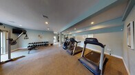 Located in St. Petersburg, Florida, this single-floor fitness center at 1175 Pinellas Point Dr S offers a well-equipped workout facility designed to support a comprehensive exercise routine. The space is finished with soft, neutral-toned carpeting and painted in a calming blue-gray palette, creating an open and inviting atmosphere throughout.

The cardio zone features multiple Cybex treadmills and an elliptical trainer, positioned along a mirrored wall to enhance the sense of space. A dedicated strength training area houses a full rack of dumbbells, a multi-functional Paramount cable machine, and a power tower for bodyweight exercises, providing options for a wide range of workout styles. Additional strength equipment, including a weight bench and stationary bike, rounds out the available amenities.

Large windows along one wall flood the space with natural light while offering views of the landscaped outdoor grounds and pool area, adding a refreshing outdoor connection to the interior environment. Recessed lighting throughout ensures consistent, even illumination for all hours of use. Decorative wall art, including colorful framed pieces and bold lettering, adds personality and visual interest to the space. An adjoining room, accessible through an interior doorway, appears to offer additional facilities. The overall layout is thoughtfully organized to allow users to move freely between equipment zones.