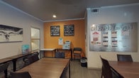 This single-floor business center is thoughtfully designed to accommodate both productivity and relaxation. The work area features multiple dark wood tables with ample seating, providing dedicated space for individual tasks or collaborative work. A printer and utility station are conveniently positioned along the vibrant orange accent wall, which is adorned with motivational typography artwork that energizes the space. A large metallic community board displays local information and announcements, adding a communal touch. Crown molding and recessed lighting lend a polished, professional finish to the room's neutral gray walls and textured carpet flooring. Adjacent to the work area is a welcoming lounge nook furnished with sleek, dark leather armchairs arranged around a side table. Large multi-pane windows framed by natural stone accent columns flood this seating area with natural light and offer direct views of an outdoor corridor, courtyard, and pool area, creating a bright and inviting atmosphere. The overall layout balances a practical work environment with comfortable gathering space, making it a versatile amenity well-suited for both focused work sessions and casual conversation.