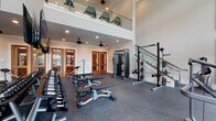 This two-story fitness facility is designed to accommodate a comprehensive range of workout activities across its two distinct levels. The ground floor serves as the primary strength training area, featuring rubber flooring throughout and an impressive double-height ceiling that creates a spacious, open atmosphere. Floor-to-ceiling windows line the walls, flooding the space with natural light and offering direct views of an outdoor pool and surrounding landscape. Equipment on this level includes a full rack of Matrix dumbbells, adjustable benches, a cable machine, a squat rack with Olympic weight plates, and various selectorized strength machines. Warm-toned wood-framed glass doors separate additional training rooms, each equipped with mirrors, wall-mounted televisions, and supplementary equipment including benches and rowing machines. Ceiling fans and recessed lighting maintain comfort throughout. A glass-railed staircase connects to the upper level, where cardio equipment—including treadmills, ellipticals, a rowing machine, and stationary bikes—is arranged along glass-panel overlooks. Large windows on the upper level provide continued outdoor views and natural light, completing a well-appointed, light-filled fitness environment.