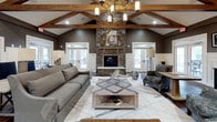 This elegantly designed single-floor space showcases a seamless blend of rustic and contemporary elements, creating a warm yet polished atmosphere. The main lounge area commands attention with dramatically vaulted ceilings accented by rich, exposed wood beams and a statement chandelier that anchors the room. A floor-to-ceiling stacked stone fireplace serves as the focal centerpiece, flanked by French doors that invite natural light and provide access to an outdoor patio area. Rich hardwood flooring in a herringbone pattern flows throughout the main areas, complementing the neutral taupe walls and crisp white wainscoting.

Comfortable seating arrangements feature plush gray sofas and intricately patterned blue accent chairs, all positioned around a geometric wood-topped coffee table. Decorative columns with classical detailing subtly define separate zones within the open layout. A dedicated business center or workroom is tucked adjacent to the main lounge, featuring a tray ceiling with a warm wood finish, built-in dark wood workstations, multiple computer terminals, and a ceiling fan. Framed artwork and carefully curated accessories, including metallic accent lamps and sculptural décor, add a layer of sophistication throughout the space.