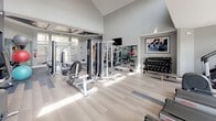 Located at 2270 Castlegate Dr N in Castle Rock, Colorado, this single-level fitness center is designed to support a comprehensive workout experience. The space is thoughtfully divided into distinct functional zones, each outfitted with professional-grade equipment to accommodate a variety of training styles.

The cardio area features a row of treadmills, an elliptical trainer, and a stair climber positioned along a wall of large floor-to-ceiling windows. These expansive windows flood the space with natural light and offer views of the surrounding community, creating an open and energizing atmosphere.

The strength training zone is anchored by soaring vaulted ceilings and a dramatic dark accent wall, giving it a bold, motivating character. This area includes a multi-station cable machine, a seated row machine, a dumbbell rack with a full range of weights, medicine balls, and wall-mounted mirrors that enhance the sense of space. Recessed and overhead lighting complements the natural light throughout.

The flooring transitions between light hardwood-style planks and durable gray tile, delineating the different workout zones. The overall design balances modern aesthetics with functional practicality, making this fitness center a well-appointed and inviting facility for daily use.