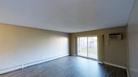This single-floor space offers a clean, functional layout with a straightforward and practical design. The main living area is characterized by dark hardwood-style flooring that runs throughout, providing a grounded, contemporary feel underfoot. Neutral-toned walls, accented by painted concrete block construction, give the space a sturdy, low-maintenance character. A smooth white ceiling completes the simple, unfussy aesthetic.

A standout feature of the living area is the large sliding glass door, which serves as both a functional entry point to an outdoor area and a generous source of natural light. The door frames a view of a landscaped green lawn and a neighboring building, creating a pleasant visual connection to the outdoors. The abundance of natural light through this opening brightens the interior and contributes to an open, airy atmosphere.

Baseboard heating is installed along the perimeter, ensuring comfortable climate control throughout the cooler months. The space is currently vacant, allowing for a clear appreciation of its dimensions and layout. Overall, the space is well-suited for those seeking a practical, easy-to-maintain environment with a connection to the outdoors and ample room to personalize with furnishings and décor.