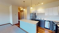 Located at 1688 Stella Avenue in Wenatchee, Washington, this single-floor residence offers a thoughtfully designed layout that balances modern finishes with comfortable living. The kitchen serves as a standout feature, showcasing dark lower cabinetry paired with crisp white upper cabinets, light quartz countertops, and a full suite of stainless steel appliances including a refrigerator, electric range, microwave, and dishwasher. A central kitchen island provides additional prep space and casual seating potential, while pendant lighting and a track light fixture add both function and style. Warm hardwood flooring flows throughout the kitchen and entry areas, creating a welcoming transition into the home. The open living area is generously proportioned, featuring plush carpeting, neutral walls, and a sliding glass door that invites natural light and outdoor access. A separate carpeted bedroom offers a quiet retreat with a large window framing views of the surrounding neighborhood and distant hillsides. The bathroom is finished with rich wood-tone vinyl flooring, a sleek white vanity with modern hardware, and a glass-enclosed shower and tub combination. Throughout the residence, clean lines, warm tones, and quality finishes come together to create a cohesive and comfortable living environment.