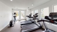 Located at 8787 Hammerly Blvd in Houston, Texas, this fitness center is a well-appointed, single-floor exercise facility designed to support an active lifestyle. The space is outfitted with multiple treadmills and an elliptical cross-trainer, providing a solid selection of cardio equipment for residents and users. The interior features clean white walls, recessed lighting, and durable carpet flooring suited for a gym environment. Large windows line multiple walls, flooding the space with an abundance of natural light and offering pleasant views of the outdoor pool area and surrounding community. The open layout allows for comfortable movement between equipment, and the bright, airy atmosphere makes for an inviting workout environment. A glass-door entry connects the fitness center to an adjacent lounge or common area, adding to the overall sense of connectivity within the facility. The combination of well-maintained equipment, ample natural light, and views of the outdoor amenities creates a functional and motivating space for daily exercise.