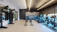 Located at 311 N Willson Ave in Bozeman, Montana, this sleek, contemporary fitness center and yoga room is a well-appointed facility designed to support a wide range of exercise routines. The space is spread across a single floor and is defined by a sophisticated modern aesthetic, featuring dark accent walls, light-toned flooring, and recessed linear LED lighting that creates a clean, energizing atmosphere throughout.

The main exercise room is generously sized and thoughtfully organized into distinct workout zones. A dedicated cardio area lines one wall, equipped with treadmills, elliptical trainers, rowing machines, and stationary bikes, all set against a dramatic hand-illustrated mountain mural — a nod to Bozeman's iconic Montana landscape. Large windows along another wall allow natural light to filter in, offering street-level views of the surrounding area. Strength training is equally well-represented, with a Torque functional trainer, kettlebell rack, Smith machine with bench press, cable machines, and resistance bands.

Adjacent to the main gym is a dedicated yoga and movement studio, featuring light hardwood flooring, floor-to-ceiling mirrors set in dark frames, and linear lighting that fosters a calm, focused environment. A water station and hallway connect the two areas seamlessly. The overall design balances performance functionality with upscale finishes, making this a premium fitness amenity.