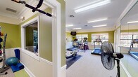 This fitness center in Daphne, Alabama offers a bright, well-organized workout environment designed to support a variety of exercise routines. The space is finished with distinctive olive-green upper walls and crisp white wainscoting, accented by crown molding that lends a polished, refined look to the athletic setting. Overhead fluorescent lighting ensures consistent illumination throughout, while multiple windows along the walls invite natural light and provide views of the surrounding greenery, creating an open and energizing atmosphere. The main workout area is generously sized, accommodating a diverse selection of cardio equipment including treadmills, an elliptical trainer, a stationary spin bike, and a rowing machine with an interactive display. A dedicated strength training alcove houses a cable machine system flanked by floor-to-ceiling mirrors, which enhance the sense of space and allow users to monitor their form. Additional large wall mirrors are positioned throughout the main floor, further amplifying the room's open feel. A wall-mounted television provides entertainment options during workouts, and a large standing fan supports air circulation. Neutral carpeted flooring covers the workout area, and direct exterior access through a glass-paneled door connects the facility to the outdoors. Posted fitness center rules indicate a community-oriented, shared-use facility maintained for the benefit of its users.
