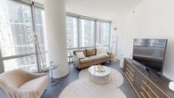 This sophisticated high-rise residence offers a seamless blend of modern design and urban living. The expansive living room is anchored by dramatic floor-to-ceiling windows that wrap around the space, flooding it with natural light and framing sweeping city skyline views. Dark hardwood flooring runs throughout, providing a rich contrast to the crisp white walls and structural columns that add an architectural dimension to the room.

The kitchen is thoughtfully designed with light wood cabinetry, stainless steel Bosch appliances including a gas range, and clean white countertops with marble-inspired backsplash detail. A pendant light and decorative gold shelving accents add a refined touch.

Both bedrooms are bright and serene, with the primary bedroom offering direct floor-to-ceiling window views of the city skyline, creating a striking visual backdrop. The rooms are finished with the same warm hardwood flooring found throughout the residence.

Two bathrooms serve the space, each finished with large-format grey tile flooring and clean white tile walls. One features a glass-enclosed shower-tub combination, while the second offers a standalone glass-enclosed walk-in shower. Light wood vanities and chrome fixtures complete the contemporary aesthetic.

A clean, white-walled hallway with dark hardwood flooring connects the private areas of the residence, maintaining a cohesive and polished design throughout.
