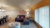 This single-floor residential unit offers a functional and comfortable layout designed for everyday living. The living room serves as the heart of the space, featuring warm accent walls, plush carpeting, and a large sliding glass door that fills the room with natural light and provides access to an outdoor balcony area. Baseboard heating runs along the walls, ensuring year-round comfort. The kitchen is efficiently designed with ample white cabinetry accented by wood-tone trim, laminate countertops, and tile flooring. It comes equipped with essential appliances including an electric range, refrigerator, and microwave, offering everything needed for home cooking. A central entryway with durable hardwood-style flooring connects the kitchen to the rest of the unit, creating a clear and organized flow. The bedrooms are carpeted and quietly appointed, each featuring windows that let in natural light and closet access. The rooms offer a restful atmosphere with neutral wall tones and textured ceilings throughout. The bathroom features a single-sink vanity with a wood-tone countertop, under-sink cabinetry, and a well-lit mirror backed by a multi-bulb fixture, providing a clean and practical space. Overall, the unit presents a straightforward, livable design with consistent finishes and a welcoming atmosphere.