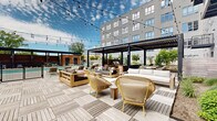 This expansive outdoor amenity courtyard is thoughtfully designed to provide a resort-like experience within a modern multi-story residential building. The space is framed by the building's contemporary grey façade, featuring large windows and private balconies that overlook the courtyard below.

At the heart of the space lies a sparkling outdoor swimming pool, bordered by smooth concrete decking and flanked by a dedicated cabana structure with lounge chairs. A gated entry ensures privacy and security for residents.

The deck area is a standout feature, finished with stylish interlocking wood tiles and sheltered by a sleek black steel pergola strung with ambient string lights. Generously arranged seating — including plush sectional sofas, accent chairs, and wooden coffee tables — creates inviting conversation zones ideal for relaxing or entertaining.

Adjacent to the deck, a lush green lawn area serves as both a recreational and leisure space, featuring a regulation bocce ball court and lounge seating. The surrounding landscaping is meticulously curated, incorporating a variety of trees, evergreen shrubs, ornamental grasses, and flowering plants set within gravel beds, adding natural texture and seasonal color. Horizontal wood privacy screens provide definition between zones while contributing warmth to the overall aesthetic. The open sky above allows for abundant natural light throughout the day.