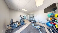 Located at 3080 E Cherry St in Springfield, Missouri, this single-floor amenity suite offers a thoughtfully organized collection of community spaces designed for both fitness and relaxation. The fitness center serves as a central highlight, featuring a comprehensive selection of True-brand cardio equipment including treadmills, an elliptical, and a stationary bike, as well as a full dumbbell rack, kettlebells, medicine balls, and a weight bench. The gym is finished with light hardwood-style flooring accented by rubber exercise mats, a dropped ceiling with recessed panel lighting, large glass sliding doors, and motivational wall graphics that contribute to an energetic atmosphere. Glass doors open to bring in natural light and views of the exterior. Adjacent to the gym is a comfortable lounge and kitchenette area, appointed with a compact refrigerator, microwave, coffee station, decorative open shelving, and casual seating arranged on a patterned area rug — ideal for unwinding before or after a workout. An office space rounds out the interior. Outdoors, a sparkling swimming pool is surrounded by a spacious patio furnished with lounge chairs, offering a refreshing retreat with open sky views and direct access from the building's exterior staircase.