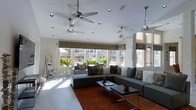 This single-floor living space offers a well-appointed, open layout centered around a generously sized L-shaped sectional sofa in charcoal gray, styled with an assortment of decorative pillows in neutral and patterned tones. A rich, burnt-orange area rug anchors the seating arrangement, adding warmth and contrast to the light-toned flooring. Two sleek, low-profile coffee tables sit at the heart of the space, providing functional surface area without obstructing the room's open feel. Multiple ceiling fans are evenly distributed across the white ceiling, complementing recessed lighting to ensure both comfort and brightness. A row of wide windows lines one wall, dressed with patterned valances in a floral motif that adds a decorative touch while allowing abundant natural light to pour in. The windows offer views of lush greenery and an outdoor area, creating a pleasant visual connection to the exterior. A slim, ladder-style shelving unit stands near one wall, adding a subtle organizational element. The overall ambiance is casual yet refined, combining practical furnishings with thoughtful design details in a comfortable, light-filled environment.