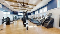 Located at 3050 Millennium Way in Kissimmee, FL, this fitness center offers a generously sized, single-floor workout facility designed to accommodate a full range of exercise needs. The space is defined by its striking architectural details, including a soaring coffered ceiling accented with exposed wood beams and oversized industrial-style ceiling fans that keep the environment comfortable throughout. Bold navy blue upper walls contrast with crisp white wainscoting below, creating an energetic yet polished atmosphere. Light wood-tone flooring runs throughout, lending warmth to the space and reflecting the abundant natural light that pours in through rows of large windows positioned along the upper walls. The open floor plan is thoughtfully organized, with a dedicated cardio zone featuring treadmills, ellipticals, stair climbers, and stationary bikes. A separate strength training area houses a comprehensive selection of cable machines, multi-use weight stations, and free weights displayed on a mirrored rack, allowing users to train across all major muscle groups. Wall-mounted televisions provide entertainment, while exercise mats and benches are conveniently accessible throughout. The mirrored walls enhance the sense of openness and support proper form during workouts. Overall, the fitness center combines functional design with an inviting aesthetic for an exceptional workout experience.